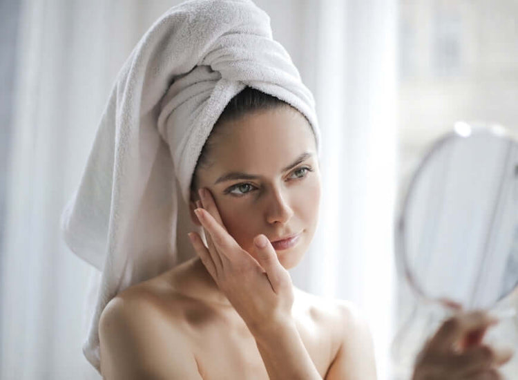 Woman with a towel wrapped around her head looking at herself in a mirror.